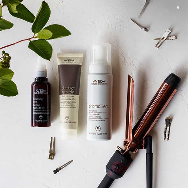 Hair care products with a flat iron, clips, and green leaves on a white surface.