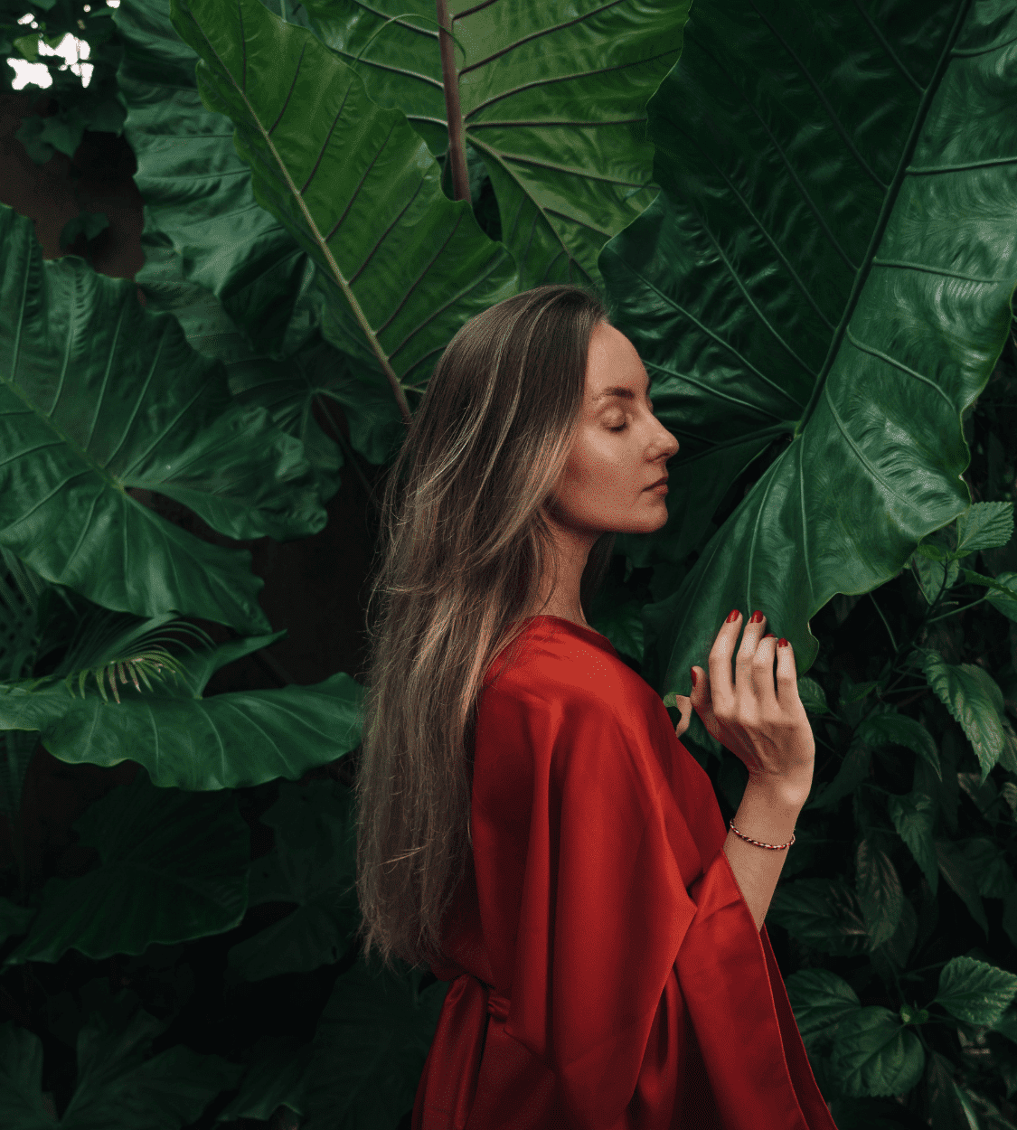 Woman in red robe stands among large green tropical leaves with eyes closed, enjoying the nature.